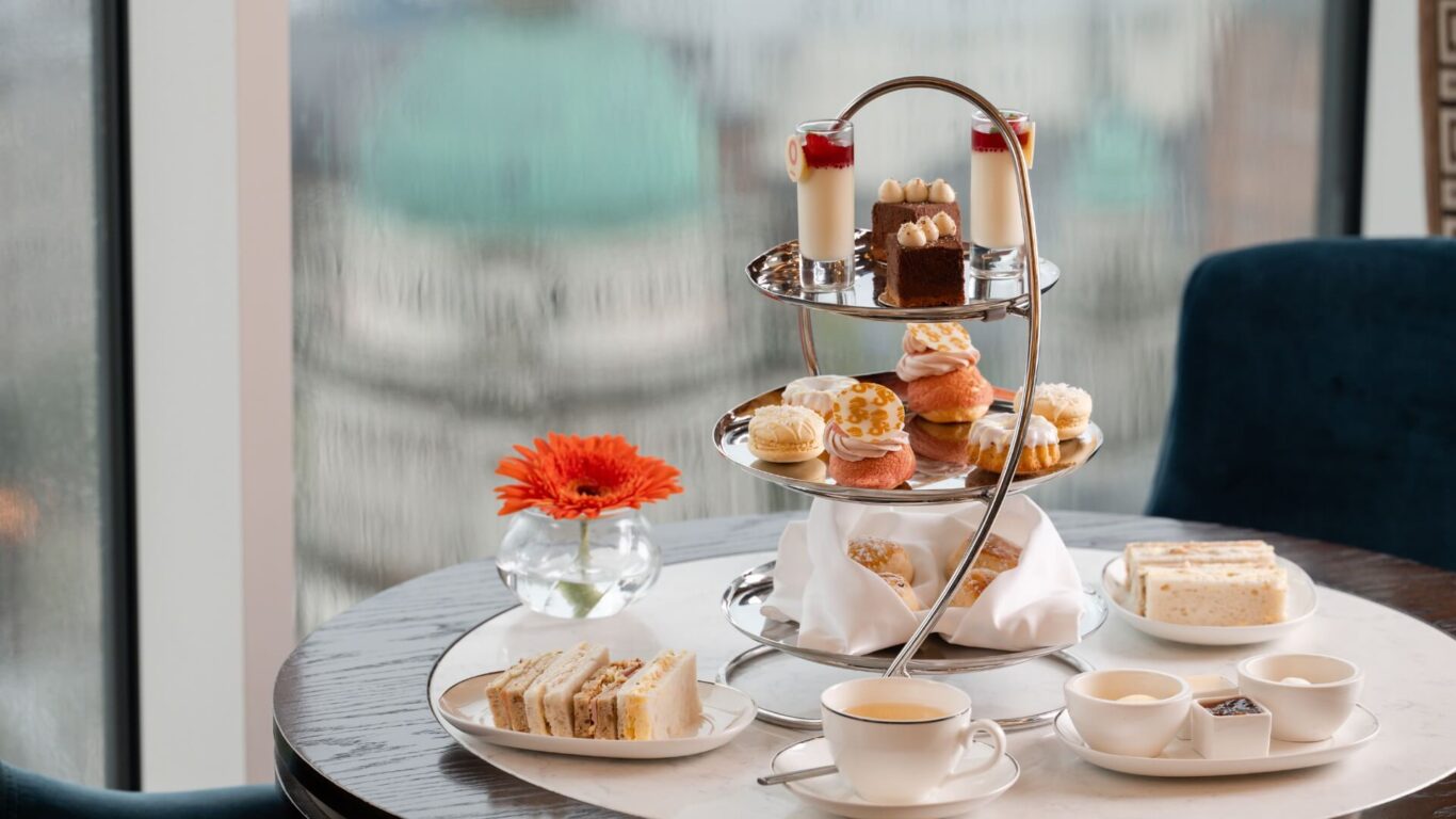 A three-tiered afternoon tea stand on a table by a window. It features assorted pastries, sandwiches, and a cup of tea.