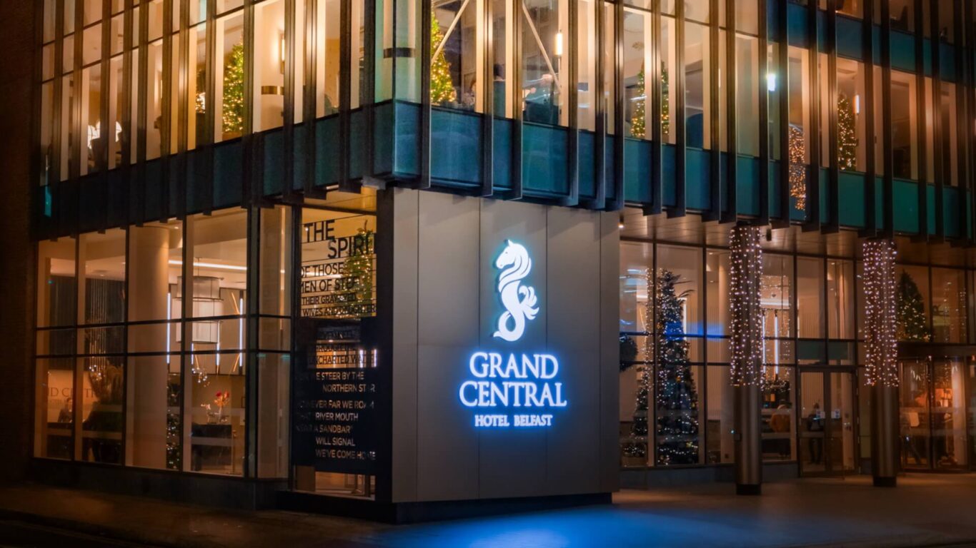 A hotel exterior at night, warmly lit with Christmas decorations. Glass windows show an elegant lobby. Bright