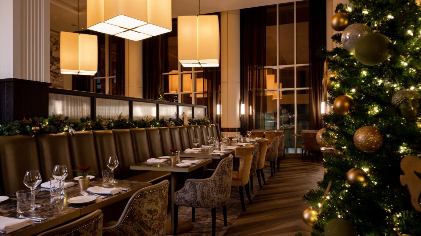 A beautiful restaurant interior with Christmas decorations. Soft lighting illuminates tables set with glassware and napkins for dining. A decorated tree glows in the corner.