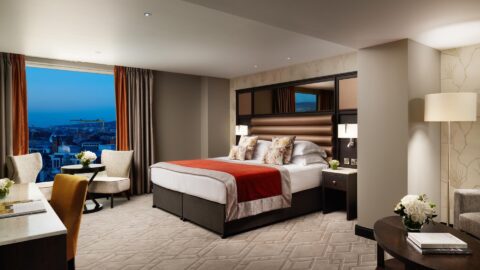 A large hotel room with a large bed, red and beige bedspread, and patterned pillows. Two chairs and a table by a window overlook a city at dusk