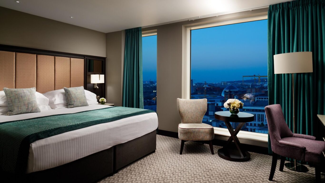 Hotel room interior with a king-sized bed covered with a teal throw. There are two chairs at a round table with flowers next to a large window with a cityscape view at dusk