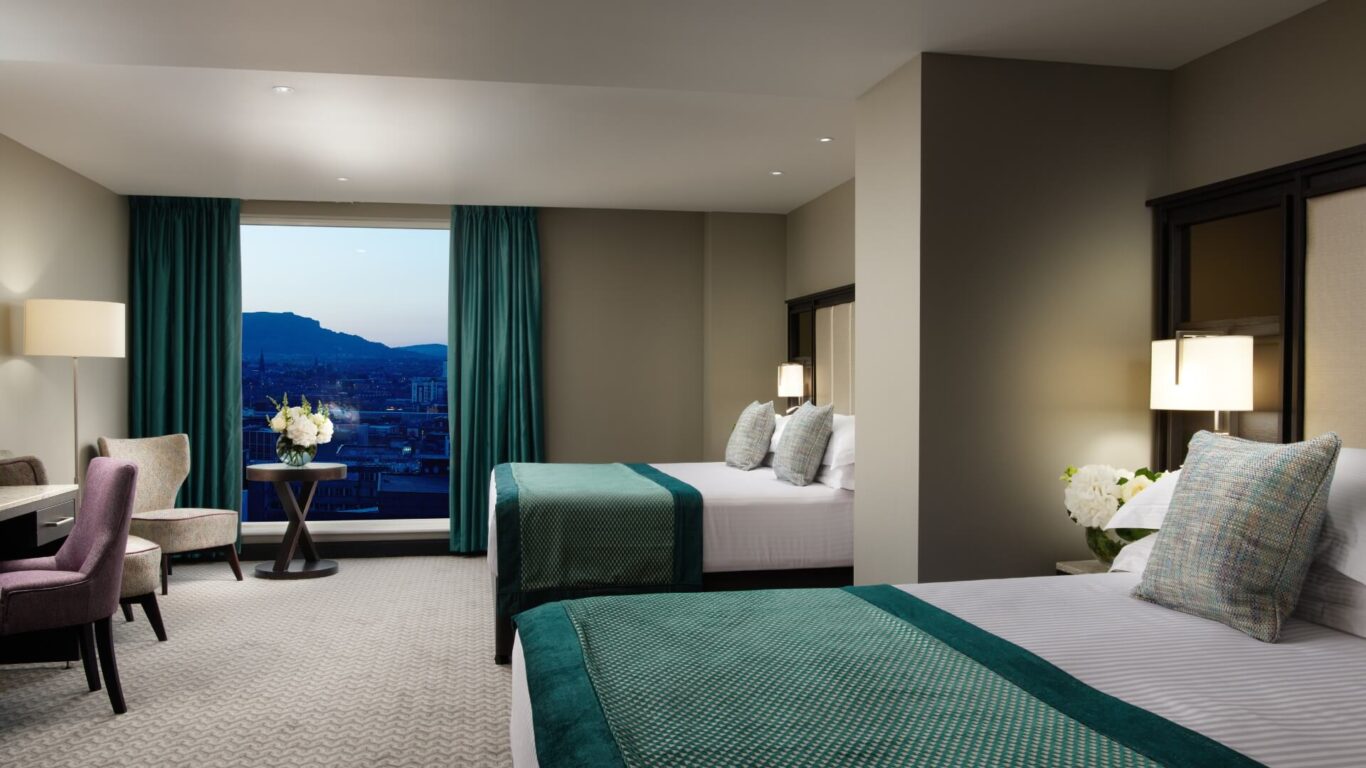 Modern hotel room featuring two double beds with teal accents, soft lighting, and a large window overlooking a cityscape at dusk.