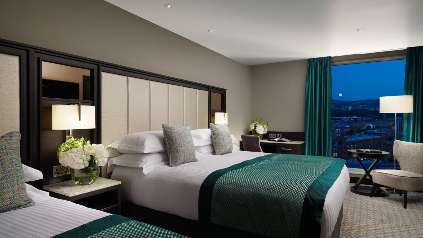 Hotel room with two double beds with teal throws. A large window shows a cityscape under a dark blue sky at dusk