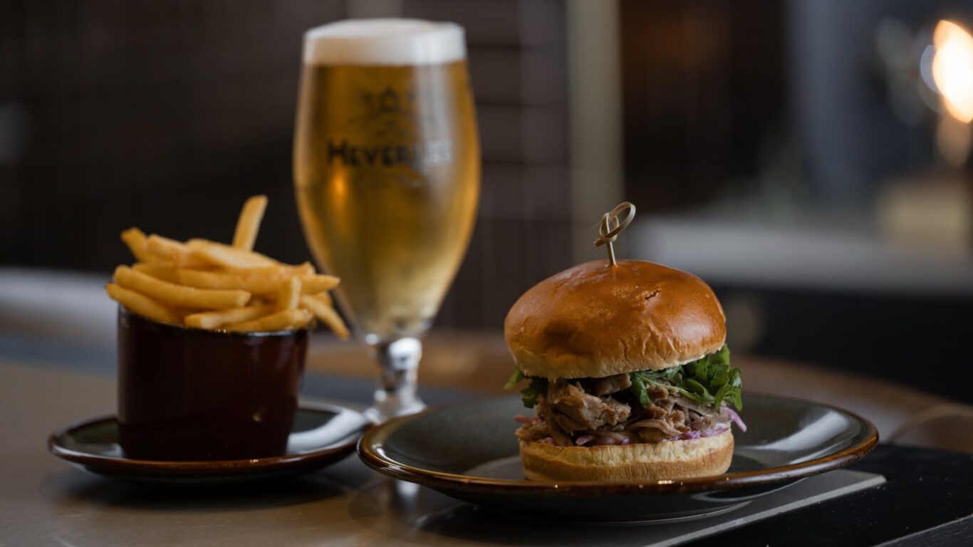 A pulled pork sandwich on a black plate next to a tall glass of beer, and chips in a cup.