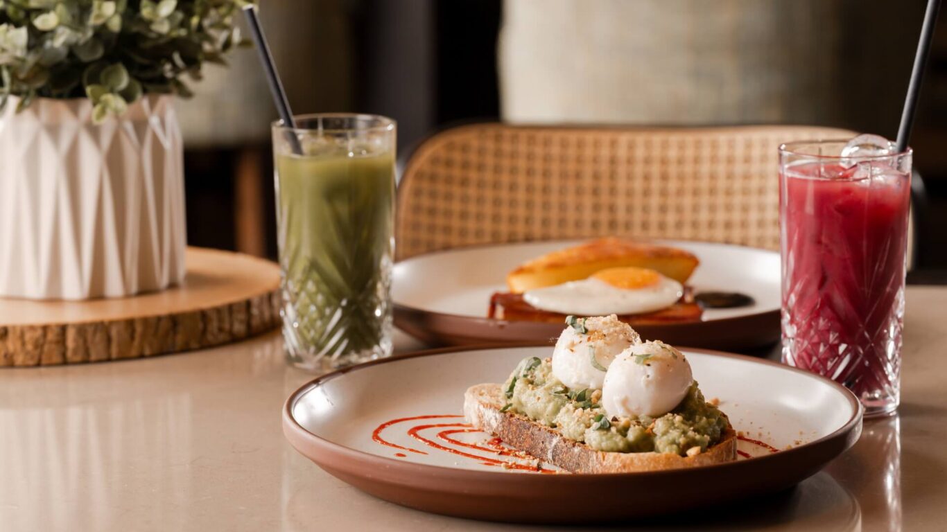Avocado toast with poached eggs on a plate sits beside a green smoothie drink. In the background, a plate with eggs and bacon, and a pink smoothie drink.