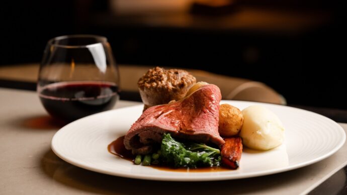 Roast beef with gravy, mashed potatoes, and roasted vegetables on a white plate, paired with a glass of red wine.