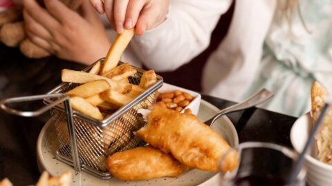 A child reached for chips in a basket next to battered fish on a plate. A small dish of baked beans sits behind the chips.
