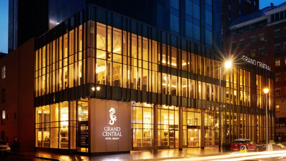 Modern hotel exterior at night, brightly lit with warm tones. Large glass windows show interior lighting. Sign reads 'Grand Central Hotel Belfast'