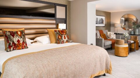 Elegant hotel room with a large bed, patterned cushions, and seating area in the background. The room consists of warm tones and modern decor