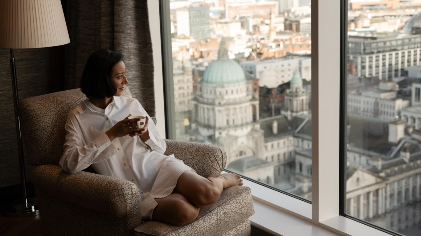 A woman in a white shirt enjoys a coffee while seated on a large chair by a window, looking down a city with historic buildings.