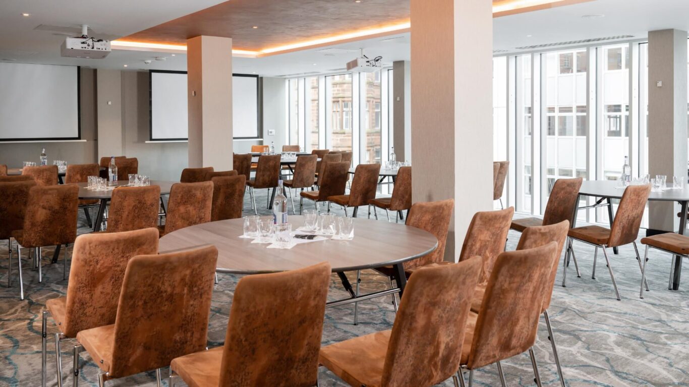 Spacious conference room with brown chairs and round tables set for a meeting. Large windows let in natural light. Two projectors and screens visible.