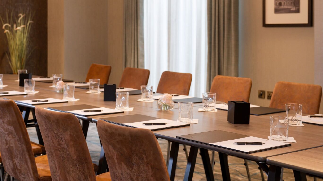 Meeting room with a long wooden table and brown leather chairs. Notepads, pens, and water glasses are neatly arranged,