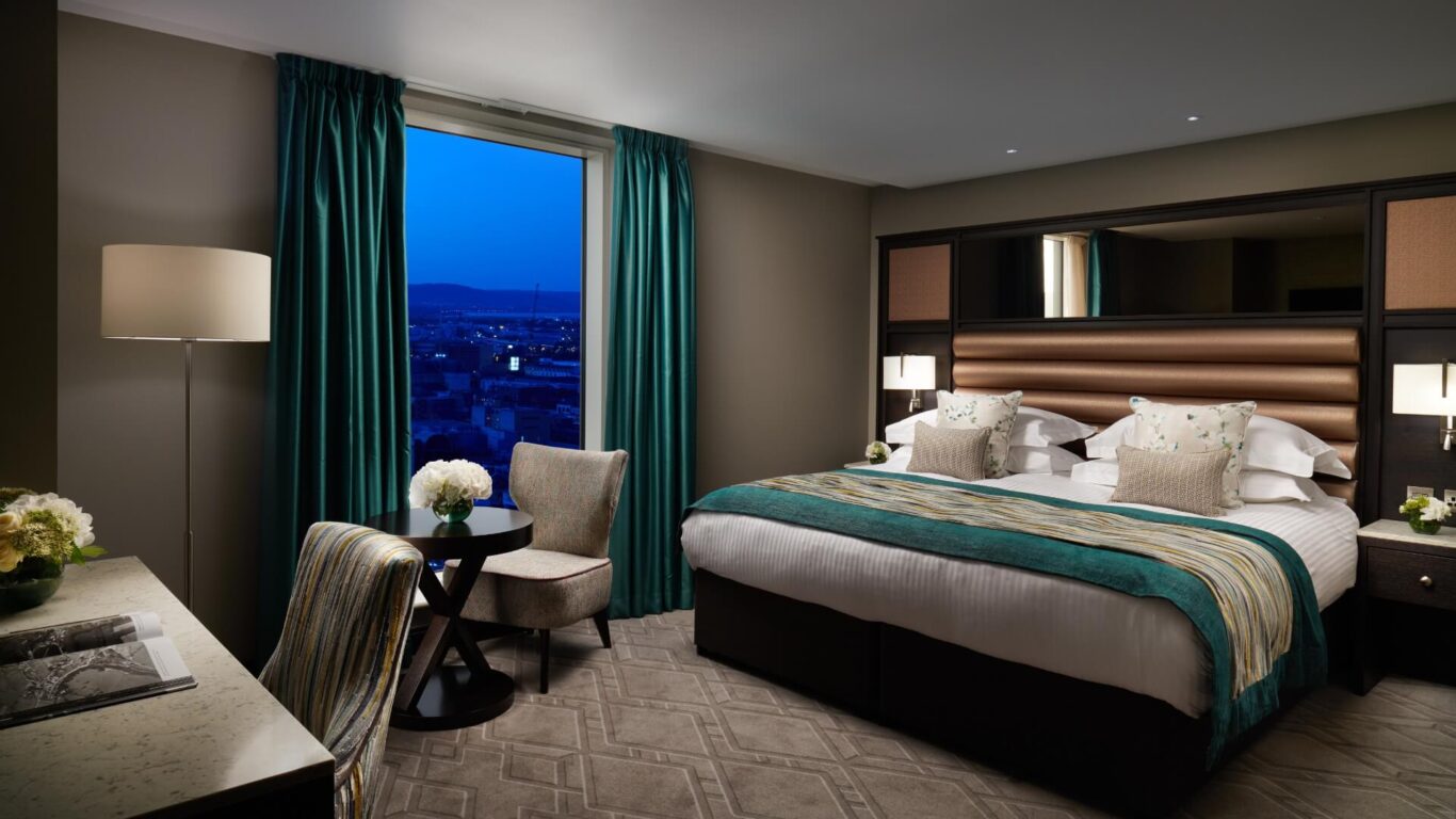 Luxurious hotel room with a king bed with teal and white bedding. A window shows a cityscape at dusk, beside which is a chair and table.