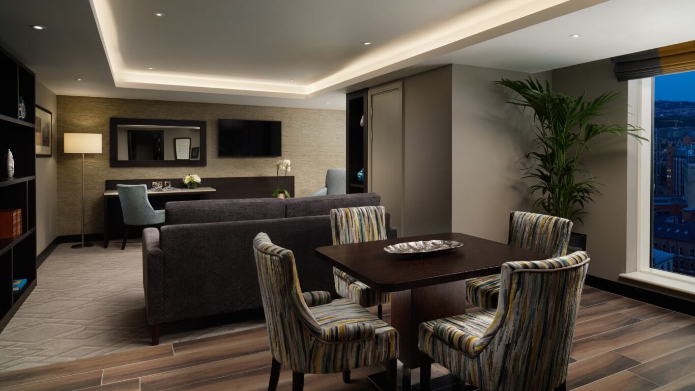 Modern living room with a dark wooden table and striped chairs sits in front of a grey sofa, which faces a TV on the wall. There is also a large window with a city view