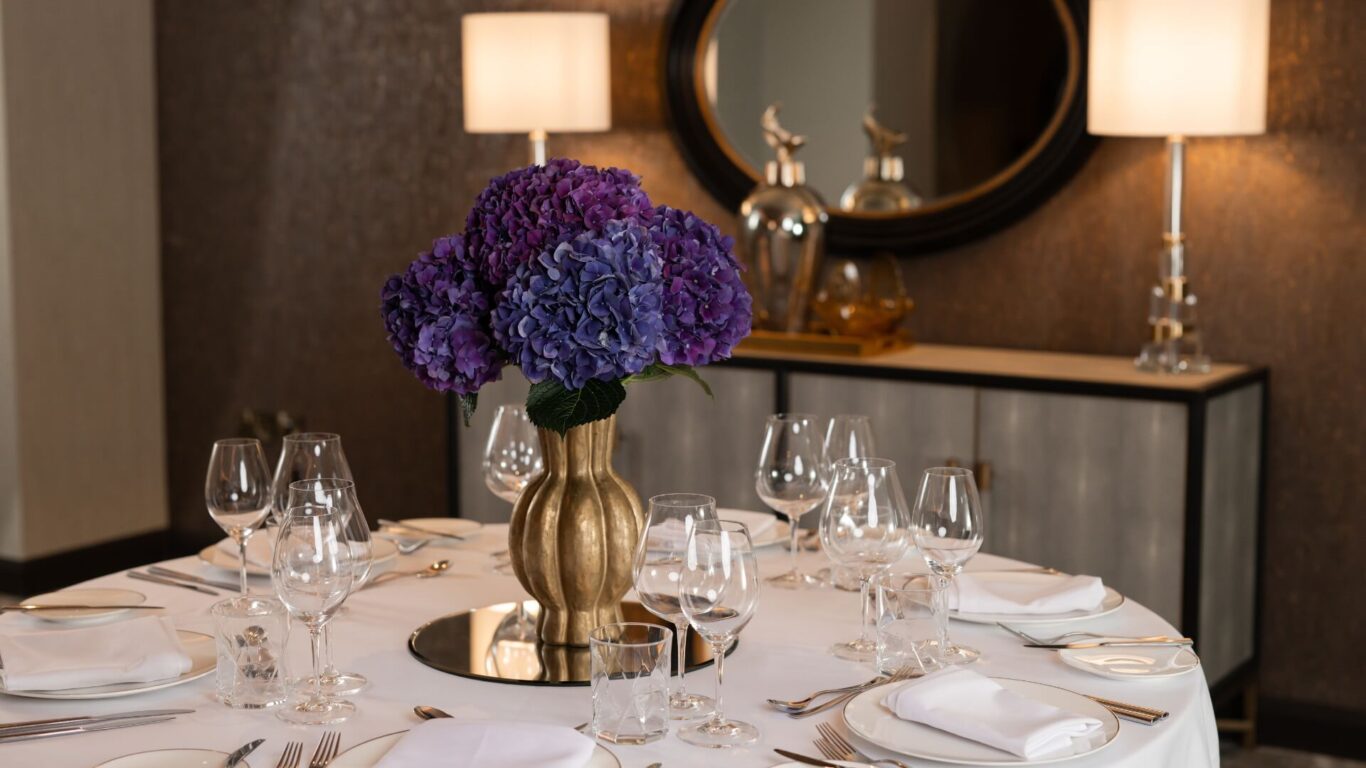 A table is set with a while tablecloth, glassware and napkins. There is a purple flower centerpiece in a golden vase