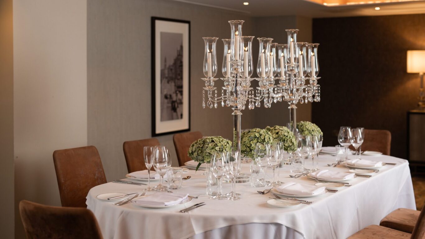 dining table set for a formal occasion, featuring a white cloth, crystal candelabra center pieces, green floral arrangements, and cushioned chairs.