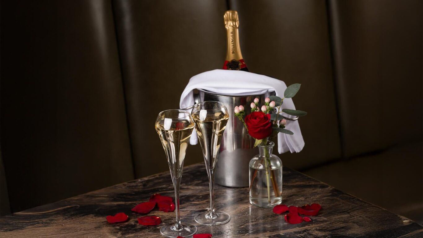 Two champagne flutes and a metal ice bucket with a bottle are on a table scattered with red rose petals and a vase with a red rose