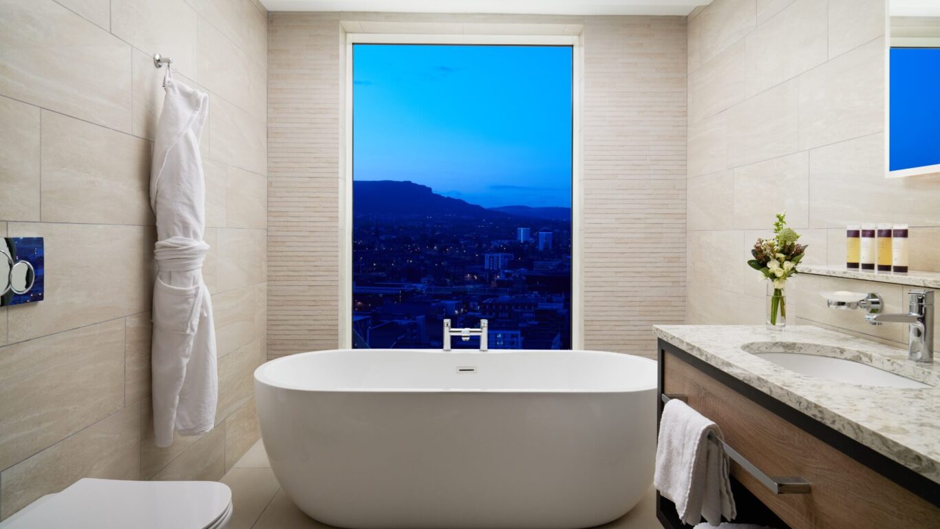 Luxurious bathroom with a freestanding bathtub, white tiled walls and a large window revealing an evening view of the city.