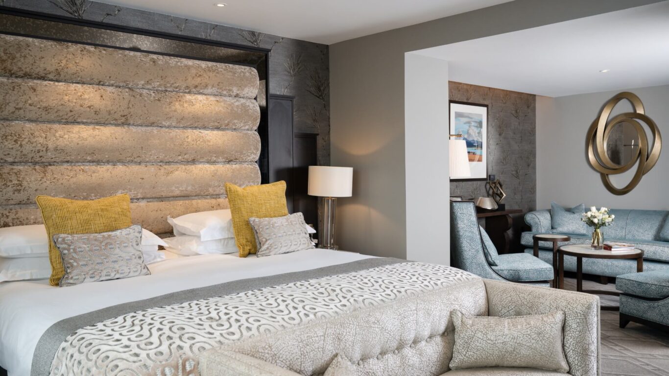Luxurious hotel room with a king sized bed with yellow and patterned pillows next to a cosy seating area with elegant décor.