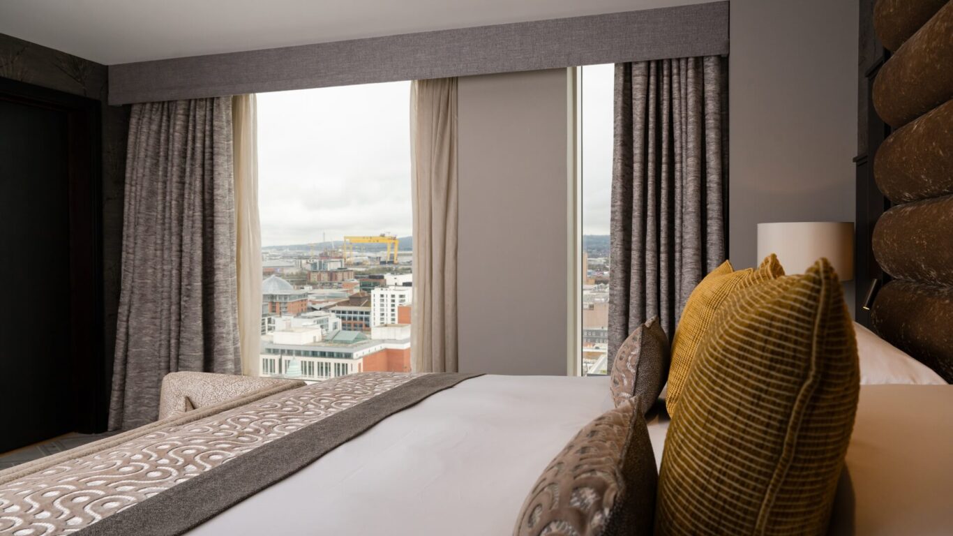 A large hotel bed facing floor to ceiling windows looking out onto a cityscape