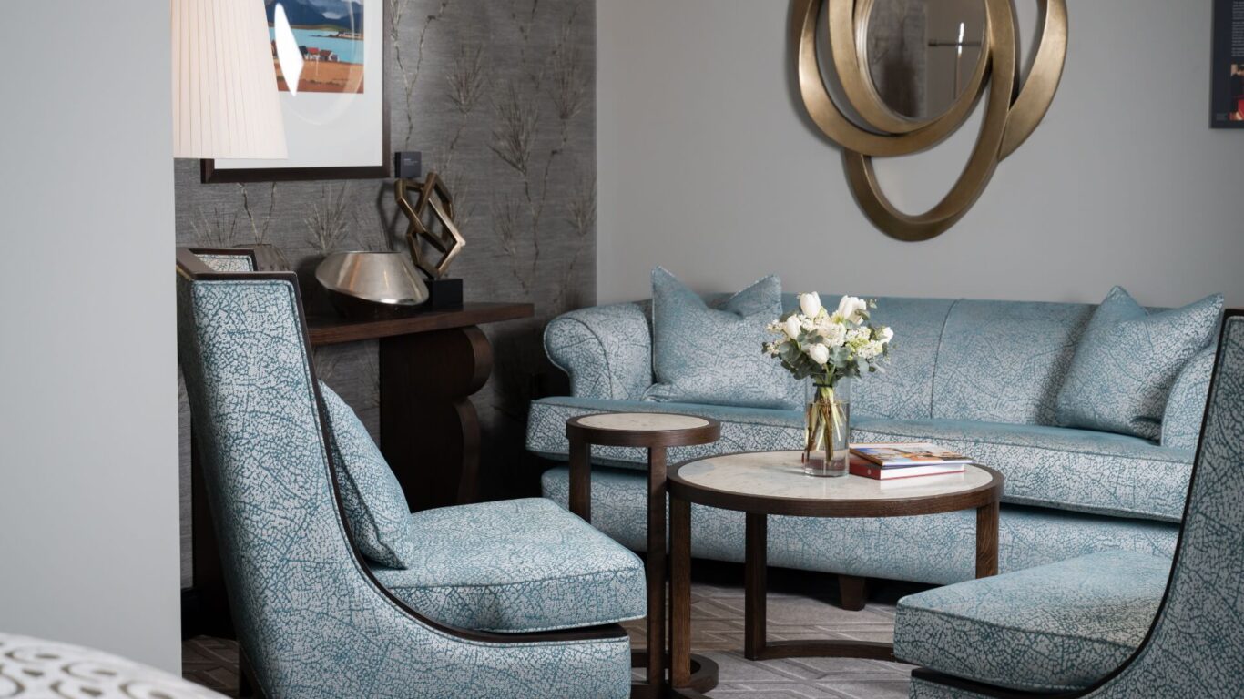 Elegant living room with teal patterned sofa and chairs, round marble-topped tables, and a decorative gold mirror on a textured grey wall.