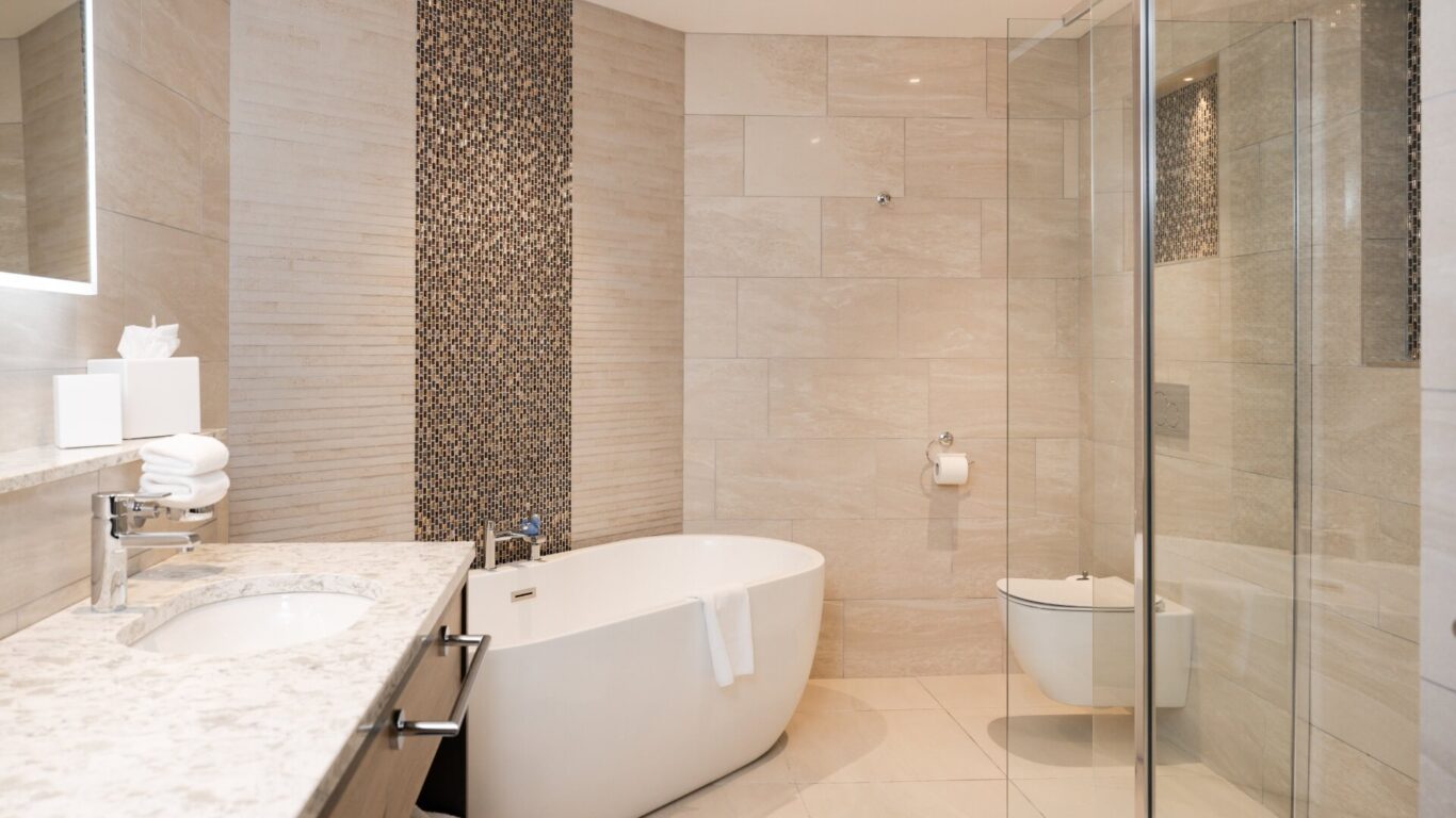 A large bathroom with a freestanding white bathtub, glass shower, and elegant marble countertop. The walls are beige tiled, with an accent wall behind the bathtub.