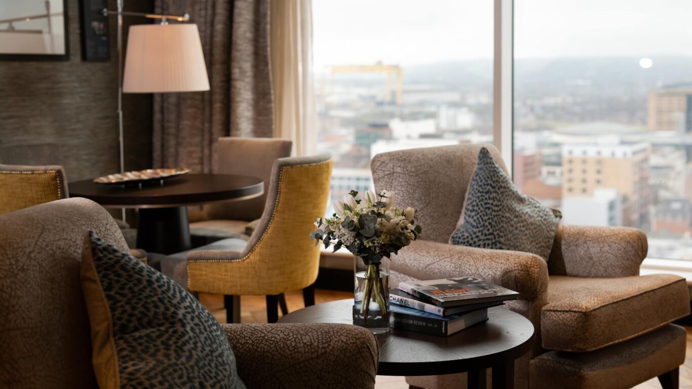 A living room space with armchairs, patterned pillows, and a round table with flowers in a vase. A large floor to ceiling window reveals a cityscape view.