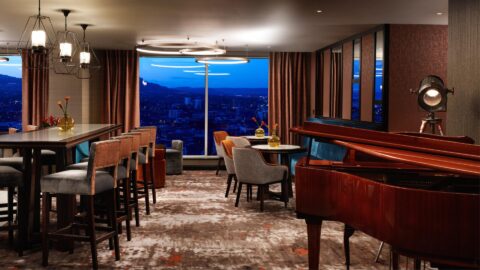 Hotel lounge with modern decor, highchairs at a long table, a grand piano, and floor-to-ceiling windows offering a cityscape view at dusk.