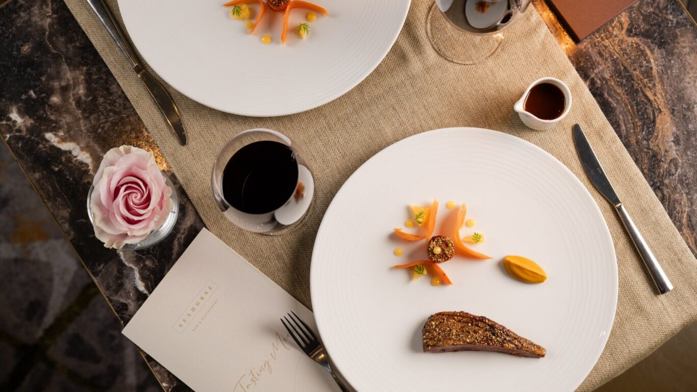 Elegant table setting with two white plates holding gourmet food artfully arranged with carrot, a meat slice, and sauce dollops. Red wine glasses next to a pink rose