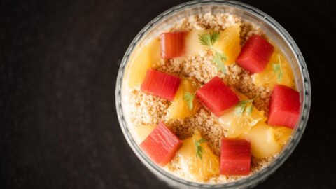 A dessert sits on a dark surface, and is topped with red rhubarb cubes, orange citrus segments, and garnished with green leaves