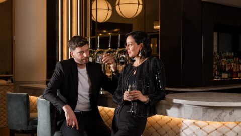 A man holding a drink and woman holding a champagne flute in formal attire lean casually against at a stylish bar.