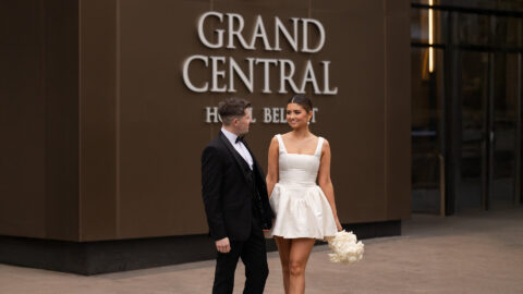 A bride and groom stand outside Grand Central hotel, dressed in wedding attire. The woman holds white flowers, and they exchange warm smiles.