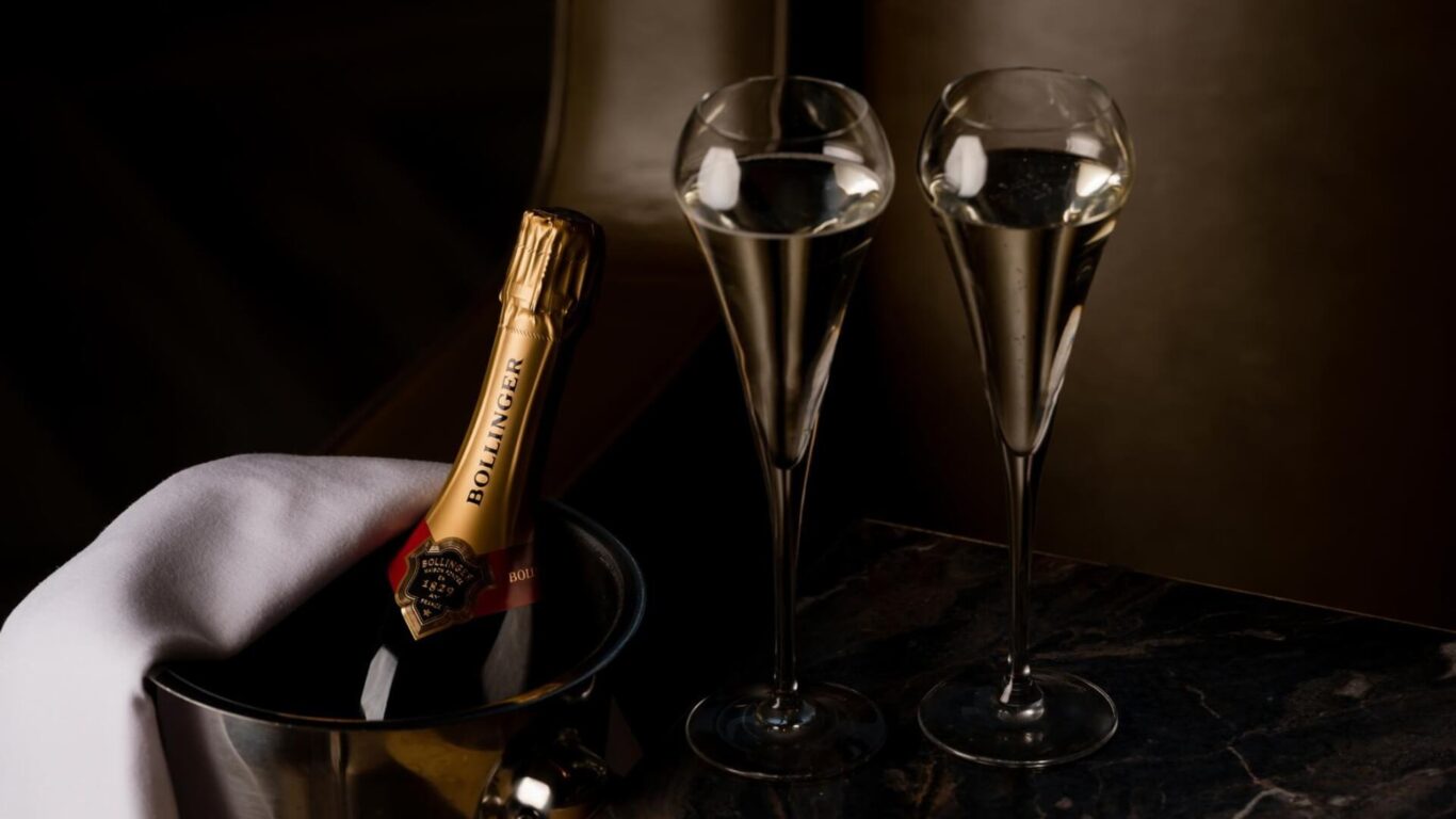A chilled champagne bottle sits in a bucket partially covered with a white napkin beside two filled champagne flutes on a dark marble table.