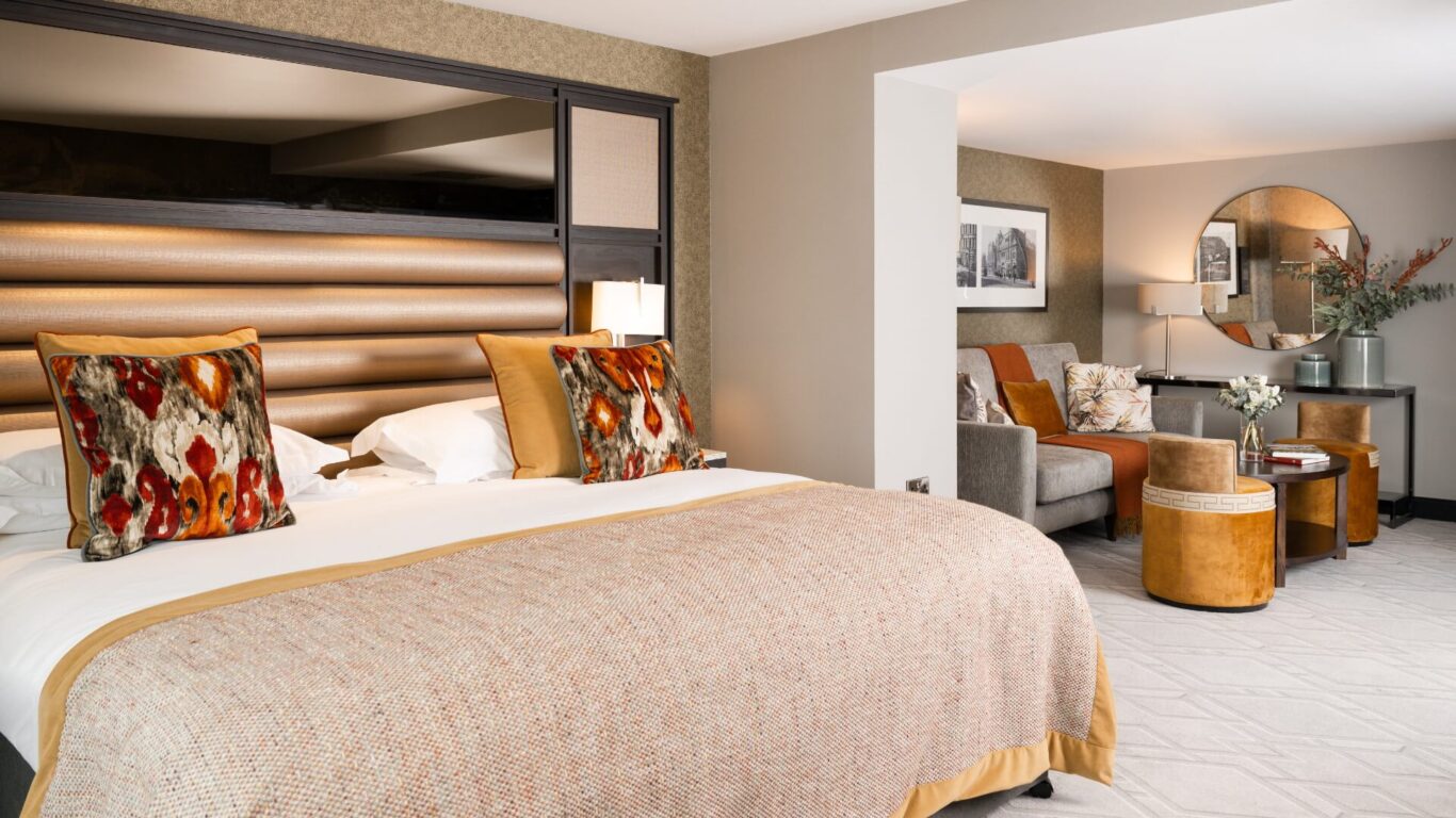 Elegant hotel room with a large bed, patterned cushions, and seating area in the background. The room consists of warm tones and modern decor