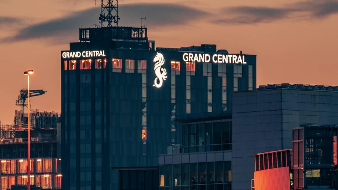 Tall building labeled with 'Grand Central' sign on a dusk cityscape, with a glowing seahorse logo against a warm toned sky.