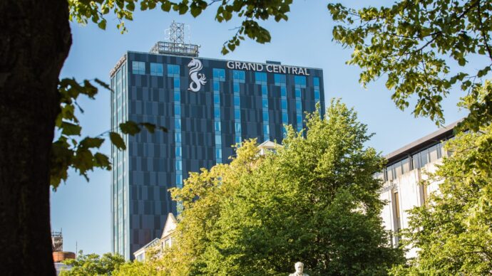 Tall modern blue building labeled 'Grand Central' in against blue skys, surrounded by vibrant green trees.