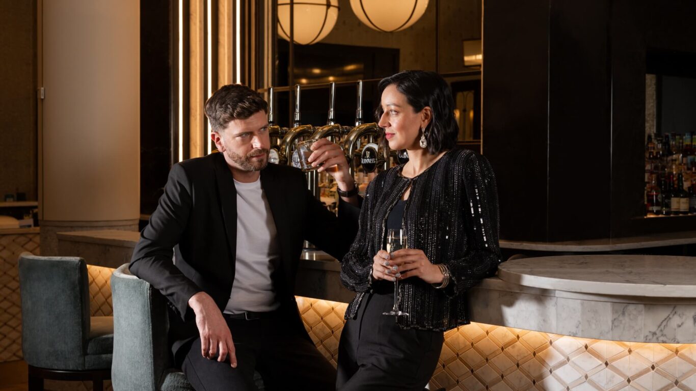 A man holding a drink and woman holding a champagne flute in formal attire lean casually against at a stylish bar.