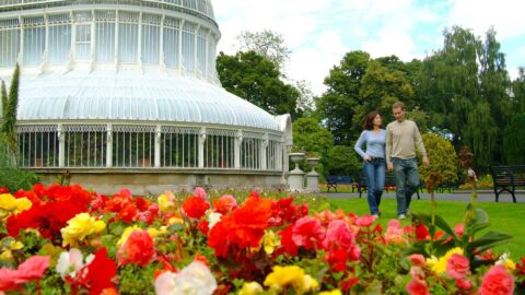 A couple strolls by a Victorian greenhouse, surrounded by vibrant red and yellow flowers, lush green trees and benches.