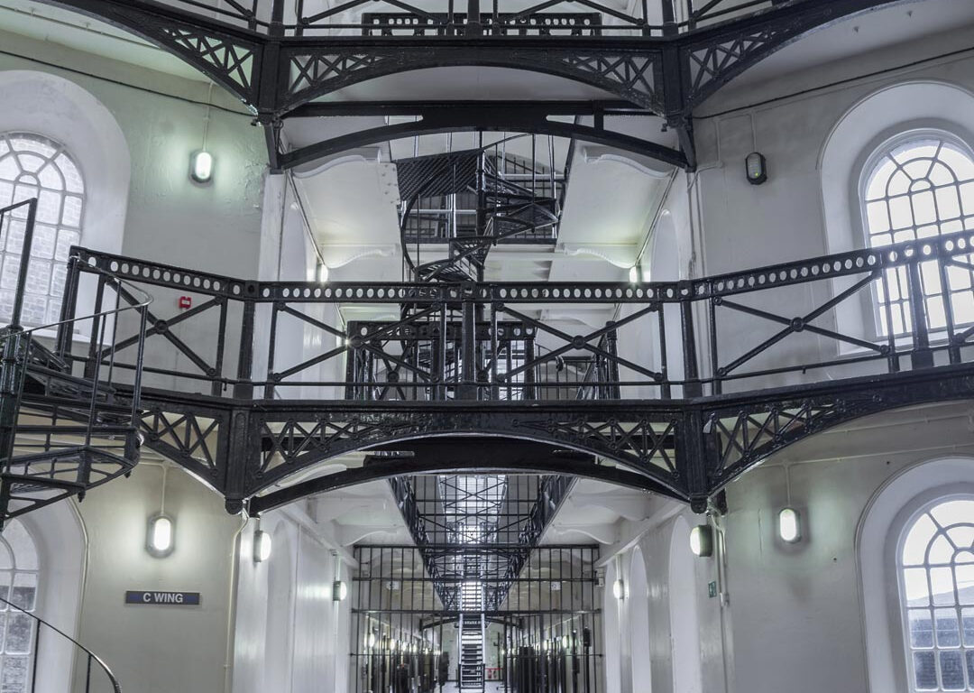 Interior of a historic building with three levels of white walls, arched windows, black metal railings and a spiral staircase
