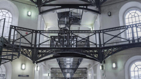 Interior of a historic building with three levels of white walls, arched windows, black metal railings and a spiral staircase