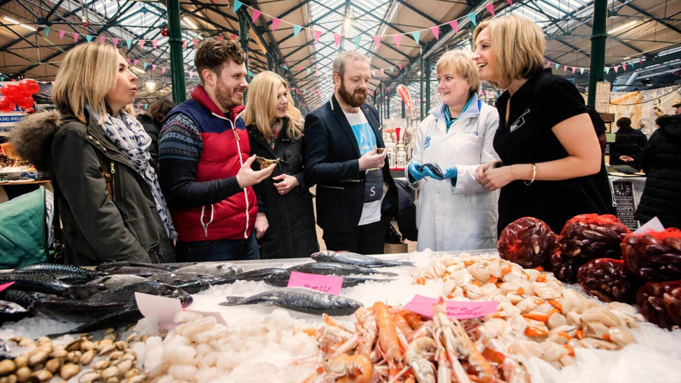 A group of smiling people stands at a market stall. A woman wearing a 