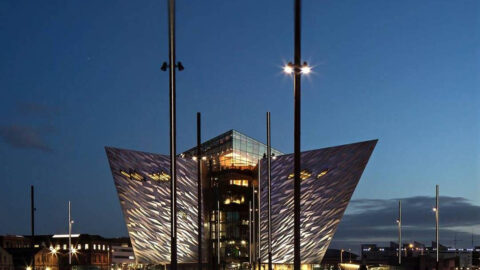 An angular, modern building with textured panels illuminated in purple and orange lights against a dark evening sky