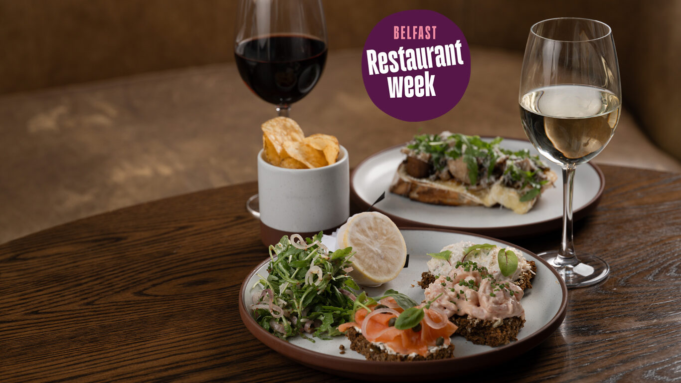 Belfast Restaurant Week Lunch dishes on a table - Annaghmore Mushroom & Ballylisk Brie Crostini and Hastings Seafood Sandwich both accompanied by a bowl of crisps and a glass of red wine and white wine